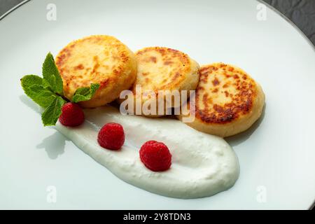 Deliziose frittelle di formaggio stese sul piatto con panna acida e lamponi decorati con menta Foto Stock