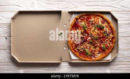 Pizza al peperoncino con peperoni, mais dolce e rucola in scatola di cartone aperta su tavolo da cucina in legno bianco con vista sul letto piatto. Spazio di copia Foto Stock