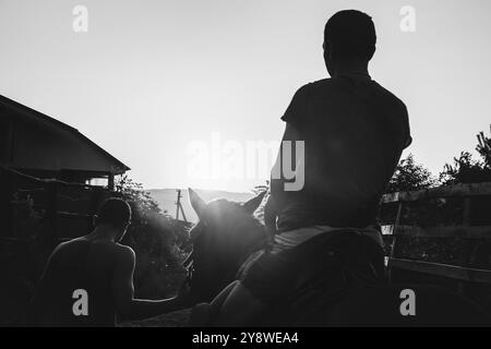 un giovane ostler conduce un cavallo vicino alla brilla con un uomo e un bambino seduti su di esso sullo sfondo di una foto estiva in bianco e nero al tramonto Foto Stock