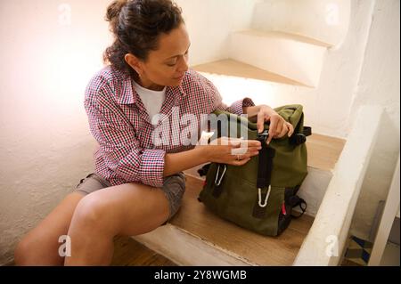 Una donna seduta sulle scale, che organizza uno zaino verde, che si prepara per il viaggio o l'avventura all'aria aperta. Atmosfera informale e concentrata. Foto Stock