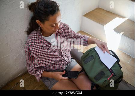 Donna seduta sulle scale, che organizza il suo zaino verde con carta e smartphone, vestita in modo informale per l'estate. Ambiente rilassante al chiuso. Foto Stock