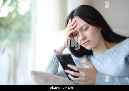 Preoccupata donna asiatica che controlla lo smartphone seduto a casa Foto Stock