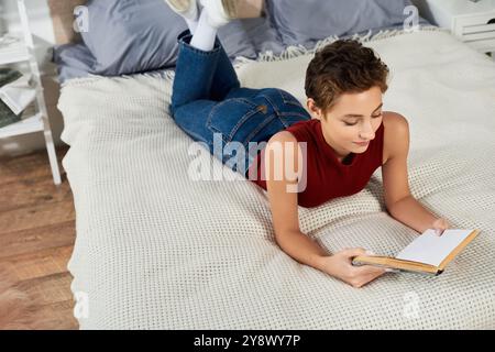 Rilassandosi a casa, una giovane donna in una canotta rossa legge un libro mentre si rilassa comodamente sul suo letto. Foto Stock