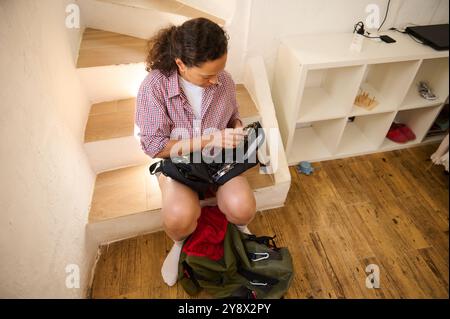 Donna che si prepara per l'escursione, seduta sulle scale, che organizza uno zaino con gli indispensabili per il trekking. Ambiente interno informale. Foto Stock