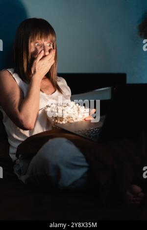 Bella donna matura seduta a letto, che mangia popcorn, scioccata e spaventata mentre guarda film spaventosi sul computer portatile. Copia spazio Foto Stock