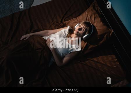 Donna stressata che ascolta la musica con le cuffie mentre cerca di dormire nel suo letto la notte. Copia spazio Foto Stock