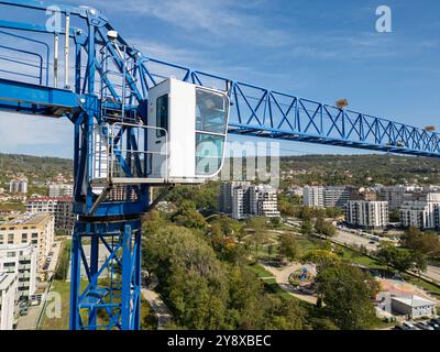 Una gru blu sovrasta un cantiere con edifici moderni e un parco sullo sfondo sotto un cielo limpido Foto Stock