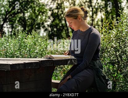 Bella donna siede tranquillamente ad un tavolo di legno, impegnata in un libro mentre è immersa nel verde, godendosi la lettura in un momento sereno nella natura estiva Foto Stock