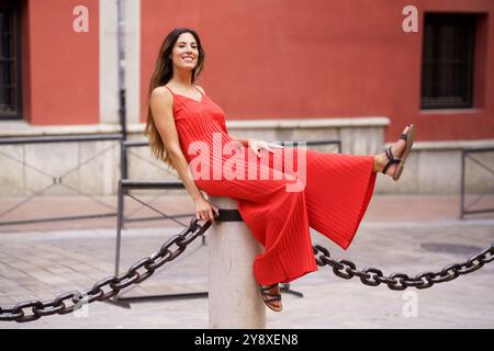 Una donna incantevole e affascinante vestita elegantemente con un abito rosso che posa giocosamente all'aperto Foto Stock