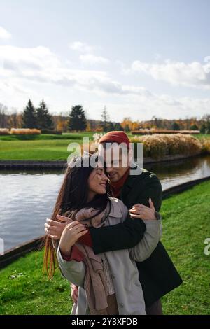 Una coppia amorevole gode di un momento tenero, avvolto in un abbigliamento autunnale, vicino a un lago tranquillo. Foto Stock