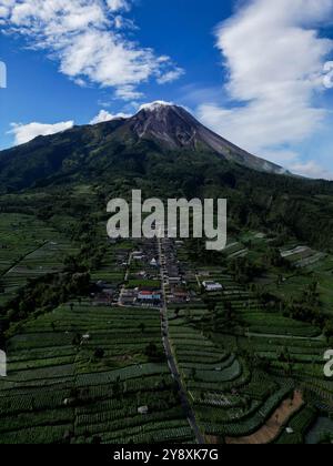 Drone verticale, le case nel villaggio di Stabelan circondate dall'agricoltura. Il maestoso Monte Merapi offre uno splendido sfondo con un cielo blu nuvoloso Foto Stock