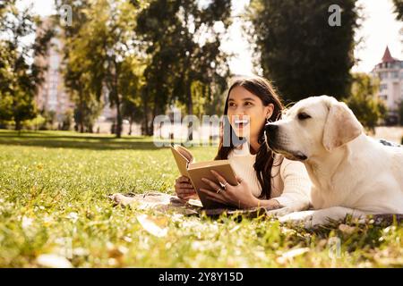 Una giovane donna in un intimo abito autunnale siede sull'erba, leggendo felicemente con il suo cane accanto a lei. Foto Stock