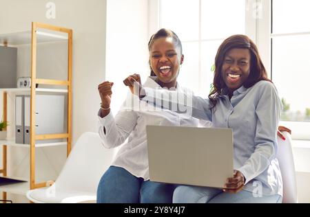 Amici africani felici e attraenti, donne d'affari che guardano un notebook, eccitati dalle buone notizie online Foto Stock