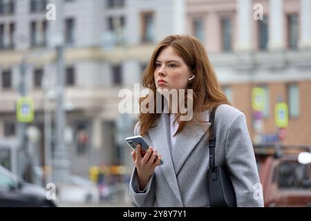 Ragazza attraente con auricolare che indossa un cappotto grigio autunnale che cammina per la città con lo smartphone in mano Foto Stock