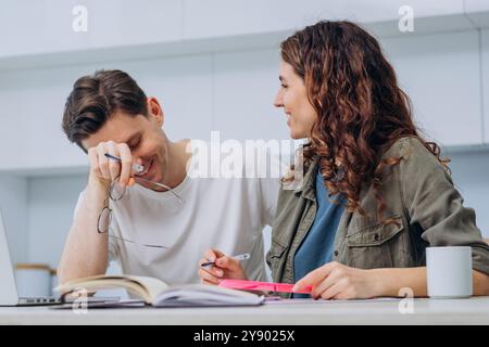 Coppia felice che lavora insieme a casa, ridendo e discutendo, lavoro di squadra informale e collaborazione. Uomo e donna sorridenti circondati da libri e notebook, godendosi momenti spensierati. Foto Stock