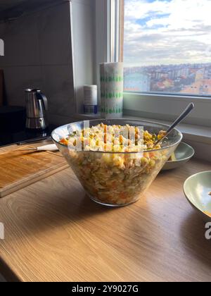 Una ciotola in vetro trasparente piena di insalata di verdure colorate si trova su un bancone della cucina illuminato dal sole. Il mix di ingredienti riflette la freschezza e la semplicità di Foto Stock