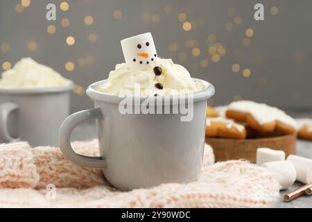 Divertente pupazzo di neve fatto con marshmallow e panna montata in una tazza di bevanda calda servita sullo sfondo contro le luci di Natale, primo piano Foto Stock