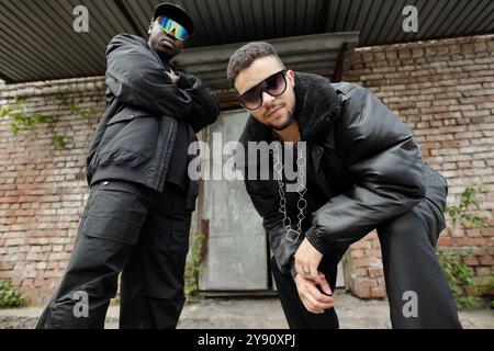 Due uomini che si pongono con fiducia in abbigliamento streetwear alla moda all'esterno di un vecchio edificio in mattoni che trasuda un'atmosfera urbana e mostra stili di moda moderni Foto Stock