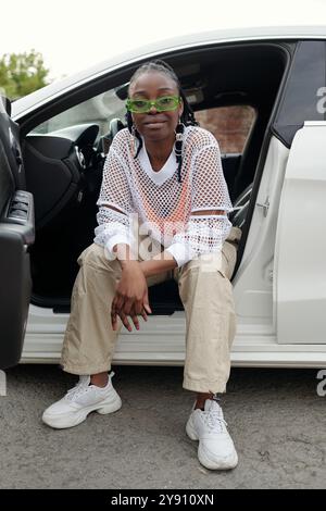 Giovane donna afroamericana con occhiali seduti sulla porta di un'auto bianca, che mostra stile casual e sicurezza guardando la macchina fotografica, parcheggiata all'aperto sul marciapiede Foto Stock