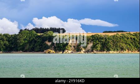 Un tranquillo paesaggio costiero in Bretagna caratterizzato da un mare turchese calmo, una scogliera verde con alberi e un cielo blu con soffici nuvole bianche. Foto Stock