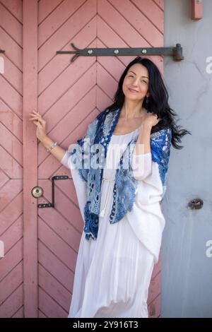 Una donna indossa elegantemente un abito bianco su una porta rustica rosa, che completa il suo scialle blu, trasudando relax e sicurezza, moda e stile di vita Foto Stock