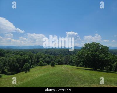 Una vista panoramica di un lussureggiante paesaggio verde con colline ondulate e montagne lontane sotto un cielo blu limpido con nuvole sparse. Foto Stock
