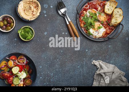 Tradizionale colazione mediorientale o brunch con uova Shakshouka in padella con toast, insalata di verdure fresche, hummus e olive su cemento rustico b Foto Stock