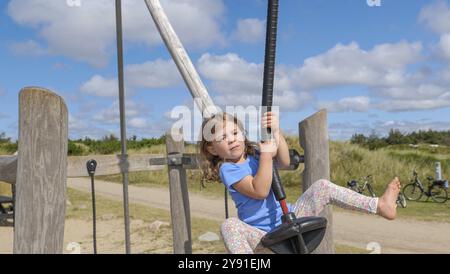 Un bambino salta felicemente su una zip wire in un ampio e soleggiato parco giochi con un cielo nuvoloso e blu Foto Stock