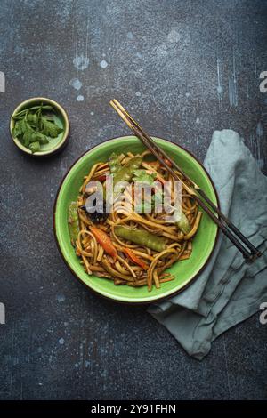 Piatto asiatico mescolare gli spaghetti di udon fritti con pollo, verdure e funghi in una ciotola di ceramica verde con bacchette di legno sul retro rustico di cemento blu scuro Foto Stock