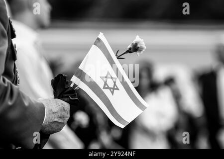 Dettagli con le mani di un uomo che regge la bandiera israeliana e un fiore durante una cerimonia commemorativa degli attacchi dell'ottobre 2023. Foto Stock