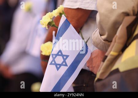 Dettagli con le mani di un uomo che regge la bandiera israeliana e un fiore durante una cerimonia commemorativa degli attacchi dell'ottobre 2023. Foto Stock