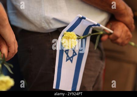 Dettagli con le mani di un uomo che regge la bandiera israeliana e un fiore durante una cerimonia commemorativa degli attacchi dell'ottobre 2023. Foto Stock