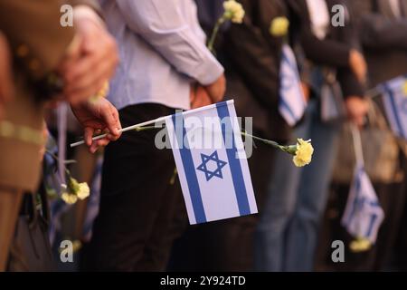 Dettagli con le mani di un uomo che regge la bandiera israeliana e un fiore durante una cerimonia commemorativa degli attacchi dell'ottobre 2023. Foto Stock
