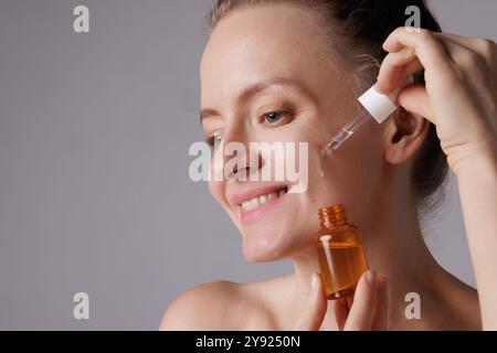 Donna sorridente felice che applica siero sul viso, occhi stretti con piacere. Bambina rossa che tiene contagocce con prodotto per la cura della pelle per sano e incandescente Foto Stock