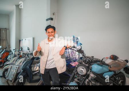 un giovane uomo vestito in modo informale mostra i pollici in piedi accanto a una motocicletta Foto Stock