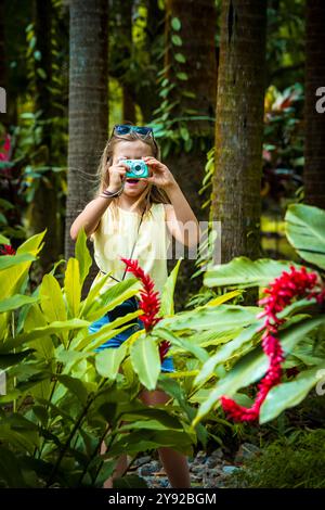 Vista di una bambina di 8 anni in piedi tra fiori di zenzero rosso in fiore con la bocca aperta a causa dell'eccitazione e per scattare foto con una fotocamera compatta Foto Stock