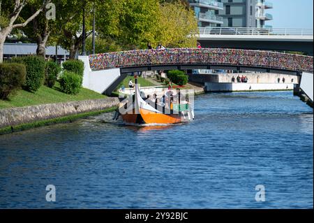 Aveiro, Portogallo - 11 settembre 2024: Una vivace barca galleggia lungo un canale mentre i visitatori ammirano il ponte adornato di chiuse. Foto Stock