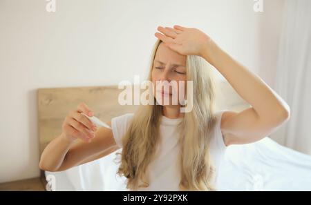 Donna matura malata che misura la temperatura corporea controllando il termometro sul letto in camera da letto di casa Foto Stock