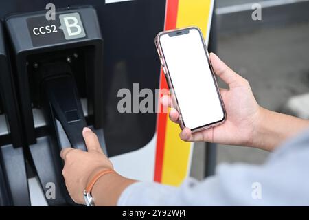 Giovane donna che usa lo smartphone mentre ricarica l'auto elettrica alla stazione di ricarica EV Foto Stock