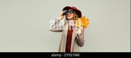 Ritratto autunnale di una giovane fotografa felice che scatta foto con la fotocamera, una ragazza elegante che tiene in mano foglie d'acero gialle che guarda lontano Foto Stock