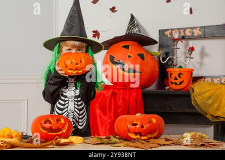 Un bambino vestito con un costume da scheletro e un cappello da strega tiene una zucca scolpita nascondendo il suo volto in modo spaventoso, con decorazioni di Halloween nel dorso Foto Stock