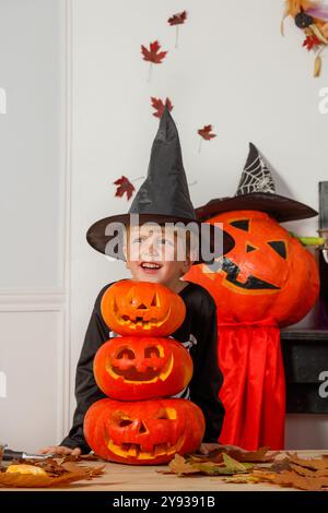 Un ragazzo sorridente con un cappello nero appuntito si siede dietro tre jack-o-lanterne, con decorazioni autunnali sullo sfondo Foto Stock