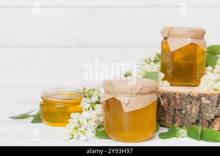 Dolce vaso di miele circondato primavera fiori di acacia. Il miele scorre da un cucchiaio in un vaso. Vasetti di miele di acacia fresco chiaro su sfondo di legno. Foto Stock
