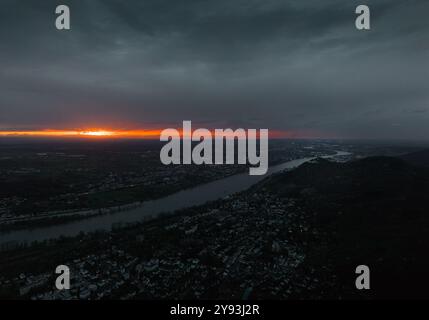 Tramonto spettacolare su un paesaggio fluviale con un vivace orizzonte arancione, nuvole scure e un vasto paesaggio urbano sottostante, creando un contrasto accattivante. Foto Stock