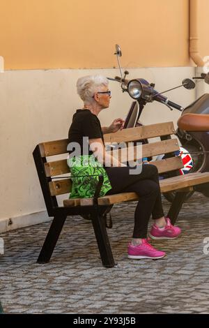 signora anziana o di mezza età con i capelli corti e grigi che indossano il nero seduto su una panchina per rilassarsi o riposarsi accanto a una borsa verde per la spesa Foto Stock