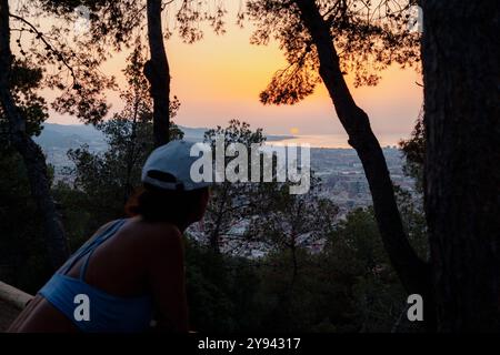 Vista posteriore di una donna irriconoscibile che ammira l'alba a Barcellona, incorniciata da alberi, con paesaggio urbano e oceano visibili in lontananza. L'orecchio tranquillo Foto Stock
