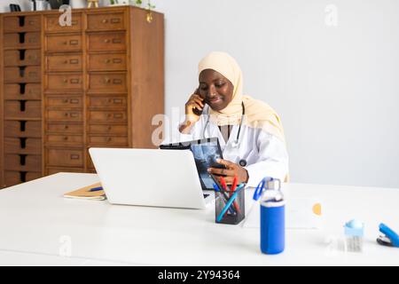 Una donna di colore professionista che indossa un hijab viene vista in multitasking in una clinica ben attrezzata, consultandosi sul suo telefono mentre si guarda la radiografia digitale Foto Stock