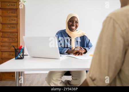 Medico africano sorridente in un hijab che si impegna con una paziente alla sua scrivania, portatile aperto e documenti pronti, in un luminoso ambiente d'ufficio Foto Stock
