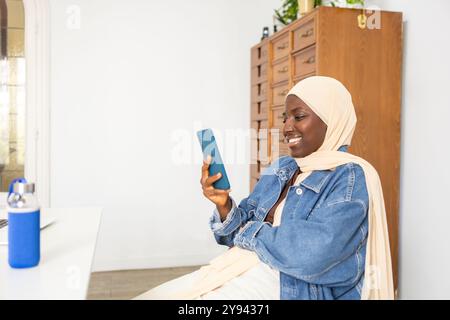 Una dottoressa nera vestita con un abbigliamento casual e indossa un hijab sorride con gioia mentre usa il suo smartphone in un ambiente d'ufficio ben illuminato Foto Stock
