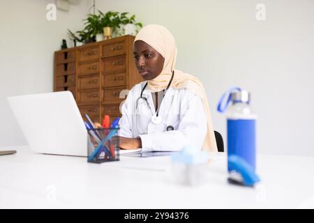 Una dottoressa nera concentrata in un hijab lavora sul suo laptop in un ufficio luminoso e moderno indossa un camice bianco da laboratorio con uno stetoscopio intorno al collo, Foto Stock